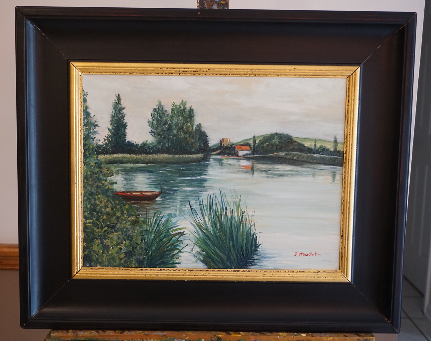 Original oil painting of a tranquil French lakeside, framed in gold and black, measuring 30 x 40 cm. Features lush greenery, a calm lake, and a small red boat, embodying a peaceful retreat for self-reflection and solace.