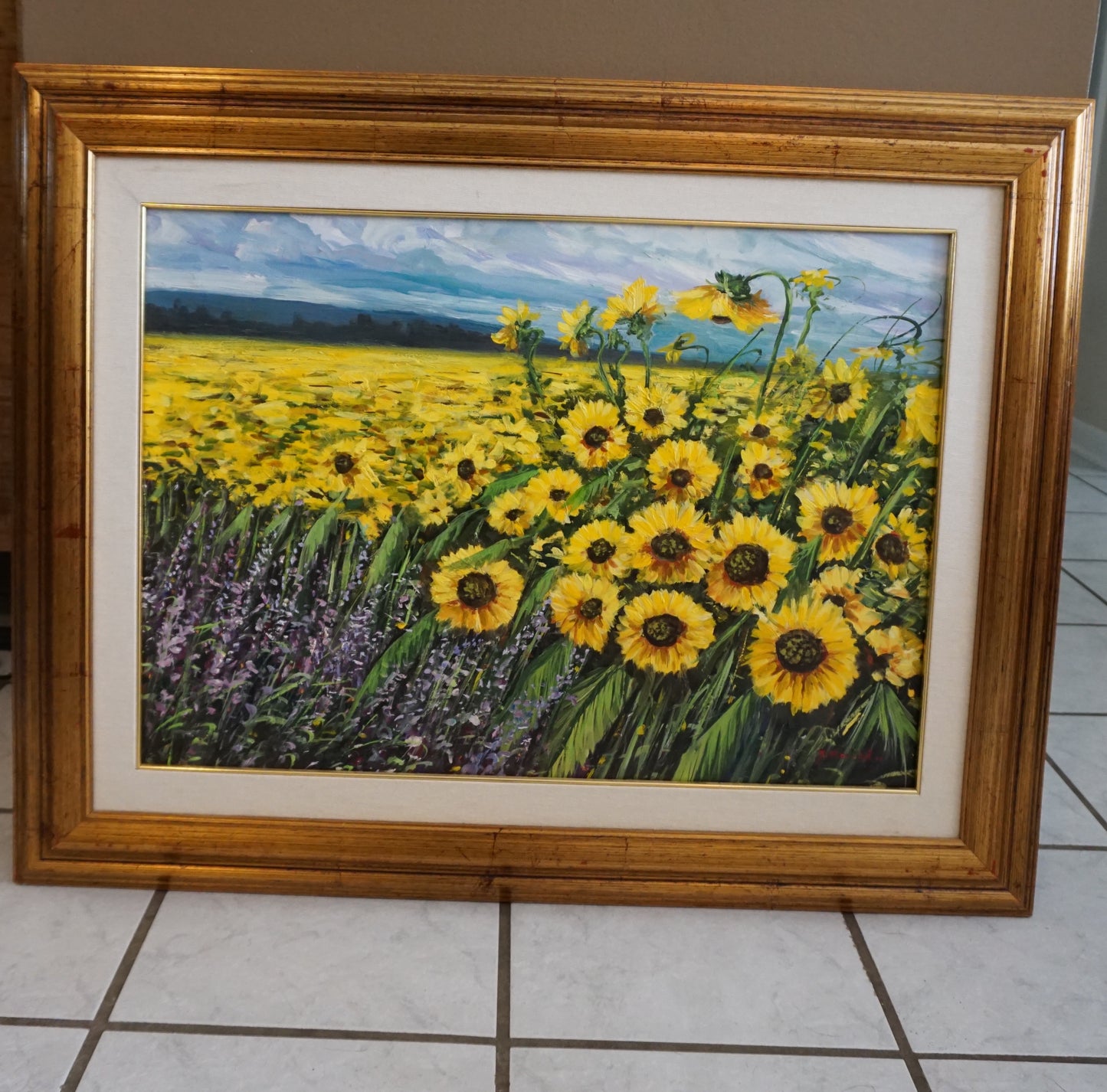 Vibrant oil painting of a vast sunflower field, with flowers in full bloom under a dynamic sky. The foreground is rich with sunflowers and lavender, set against a backdrop of distant mountains, all encased in a golden wooden frame. This piece evokes the warmth and energy of a sunny day in nature.