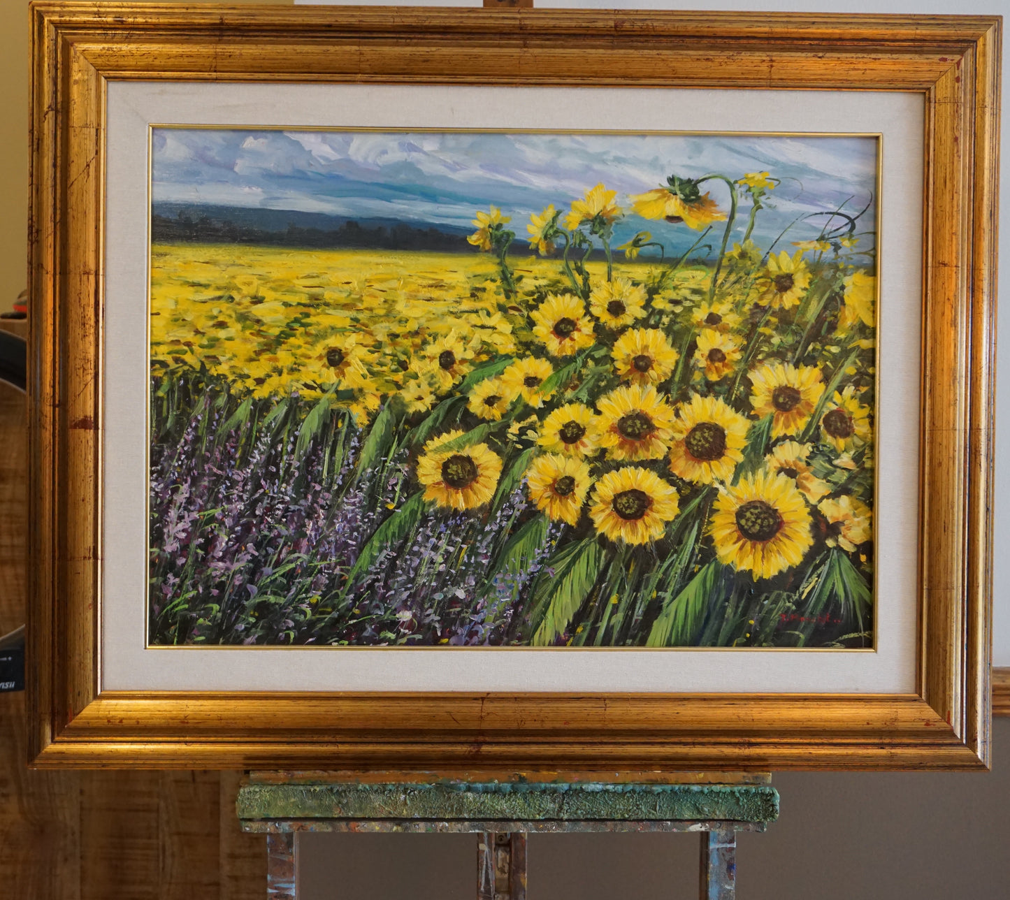 Vibrant oil painting of a vast sunflower field, with flowers in full bloom under a dynamic sky. The foreground is rich with sunflowers and lavender, set against a backdrop of distant mountains, all encased in a golden wooden frame. This piece evokes the warmth and energy of a sunny day in nature.